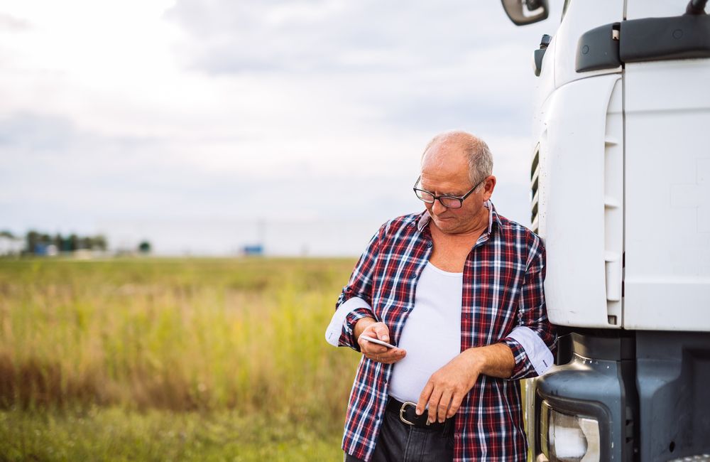 Qual o perfil de um caminhoneiro? Na imagem, transportador ao lado do seu caminhão de transporte lendo sobre o assunto em seu celular.